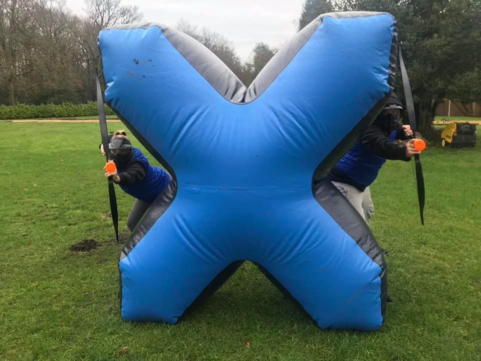 Two players aiming around a blue X-shaped inflatable bunker wearing protective face masks