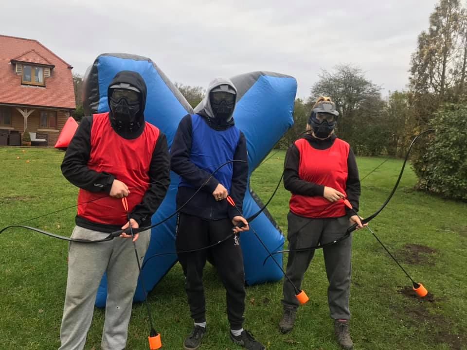 Three players holding bows with foam-tipped arrows in front of an inflatable X-shaped barrier