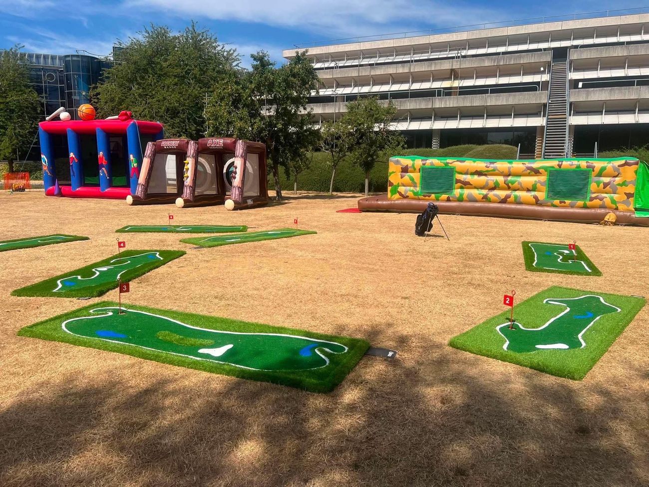 Portable putting course with numbered holes on a corporate campus lawn, golf bag beside it
