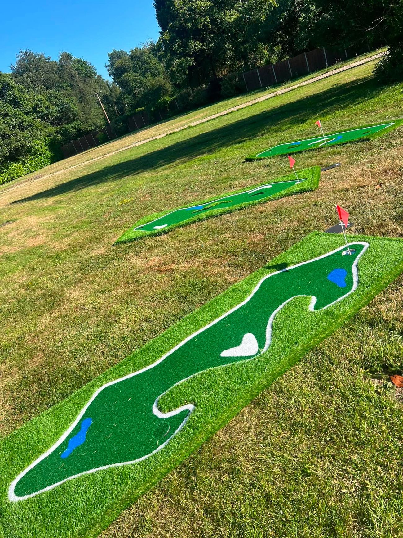 Three winding raised mini golf holes across a summer lawn, trees and blue sky behind