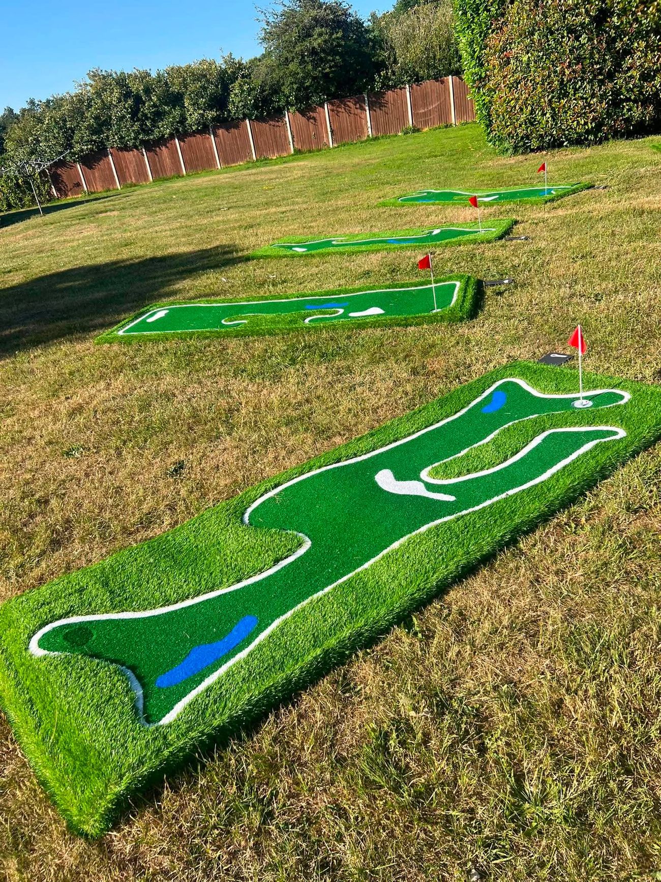 Four raised mini golf holes set diagonally up a lawn with a wooden fence behind