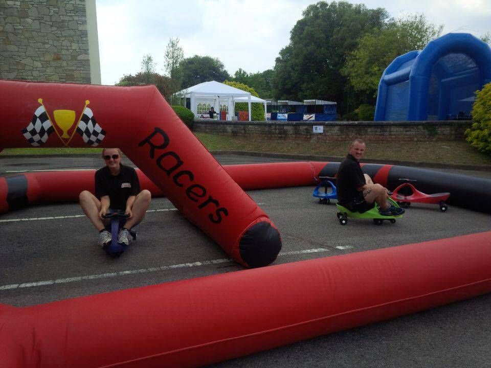 Two people sitting on Didi cars inside the red inflatable Racers track on tarmac, a bouncy castle behind