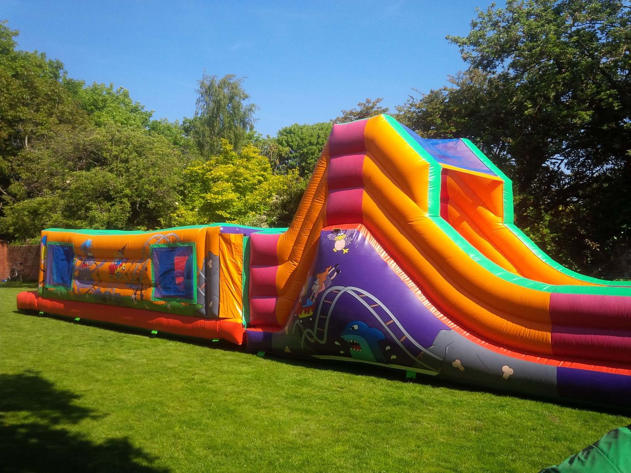Wide side view of full course on grass, obstacle section left, tall slide with purple artwork right
