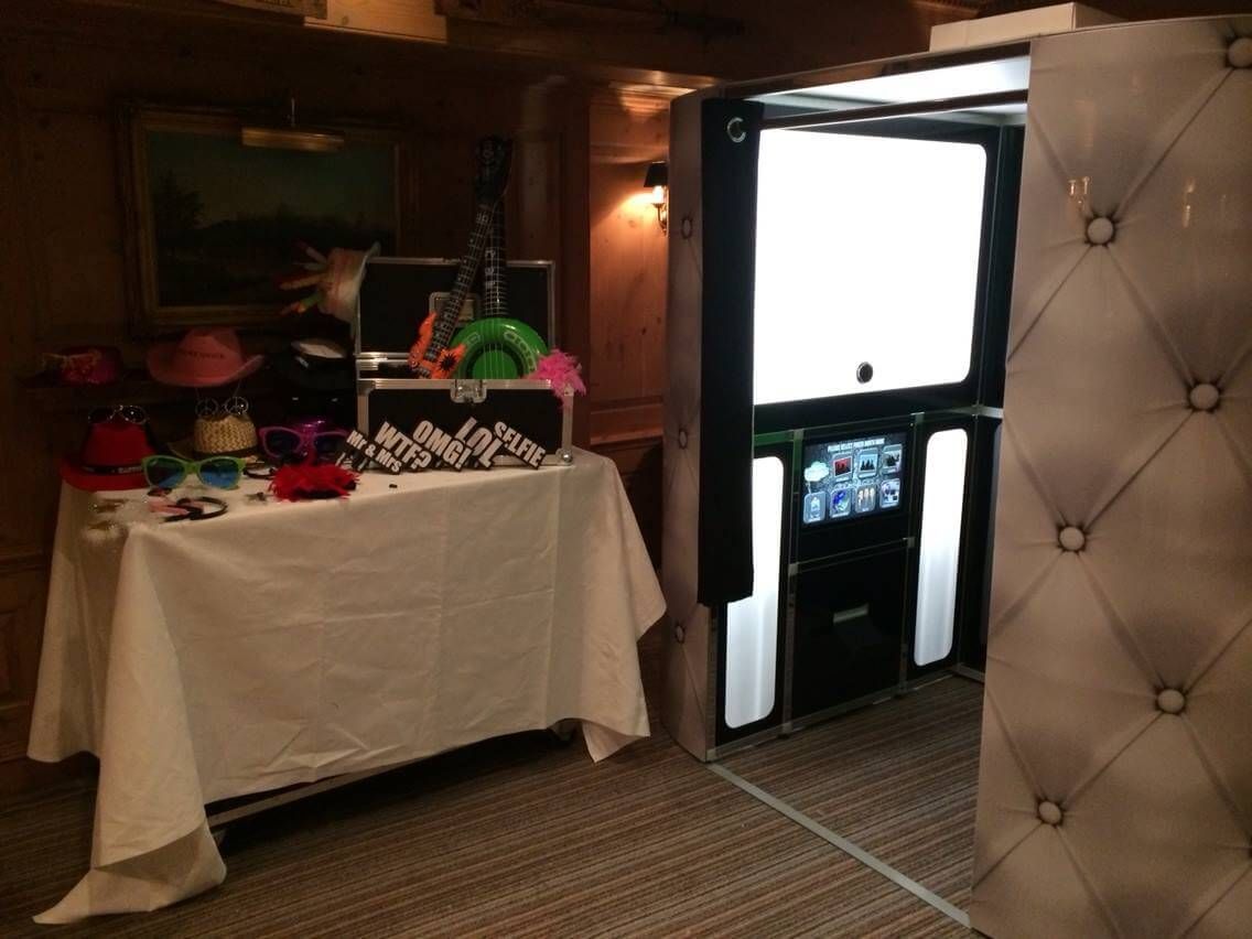 Silver tufted photo booth set up indoors beside a table of hats, glasses and props