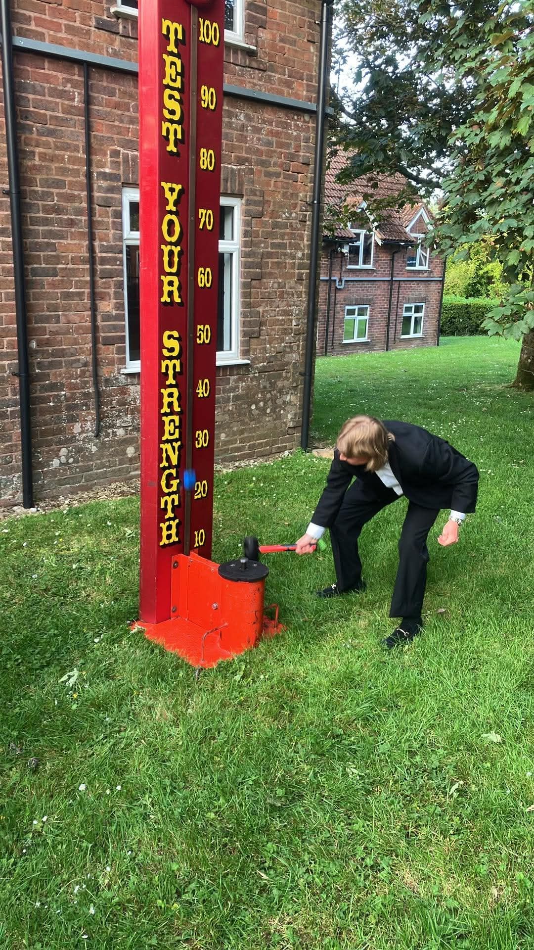 Test Your Strength high striker set up on grass, with a player swinging the mallet at the base and the numbered scoring strip running up to the bell