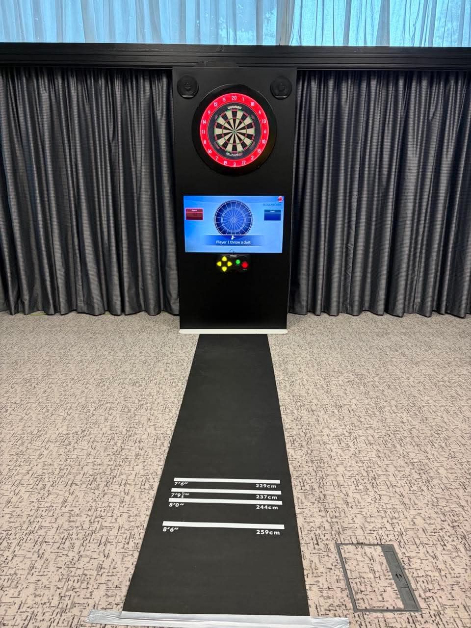 Black auto-scoring dartboard cabinet with lit board surround and score screen behind its throw mat