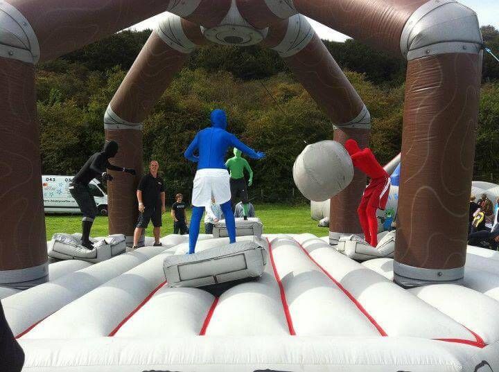 Four players in coloured morphsuits on the pedestals swinging at the central padded ball