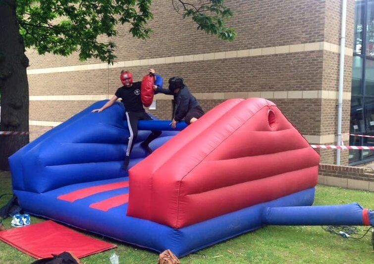 Two players in red and black head guards balancing on the pole of the jousting inflatable, brick building behind