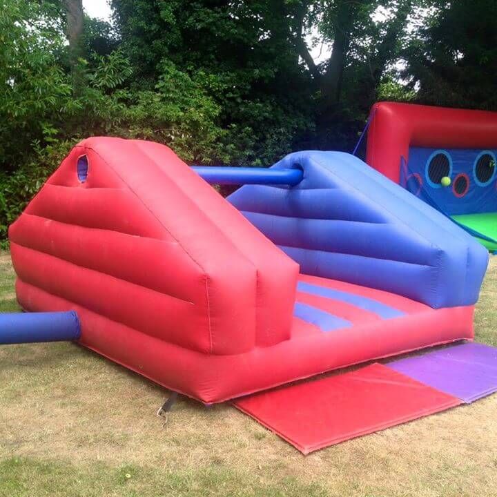 Empty red and blue pole jousting inflatable with a central pole and padded ends, set up on grass beside a hedge