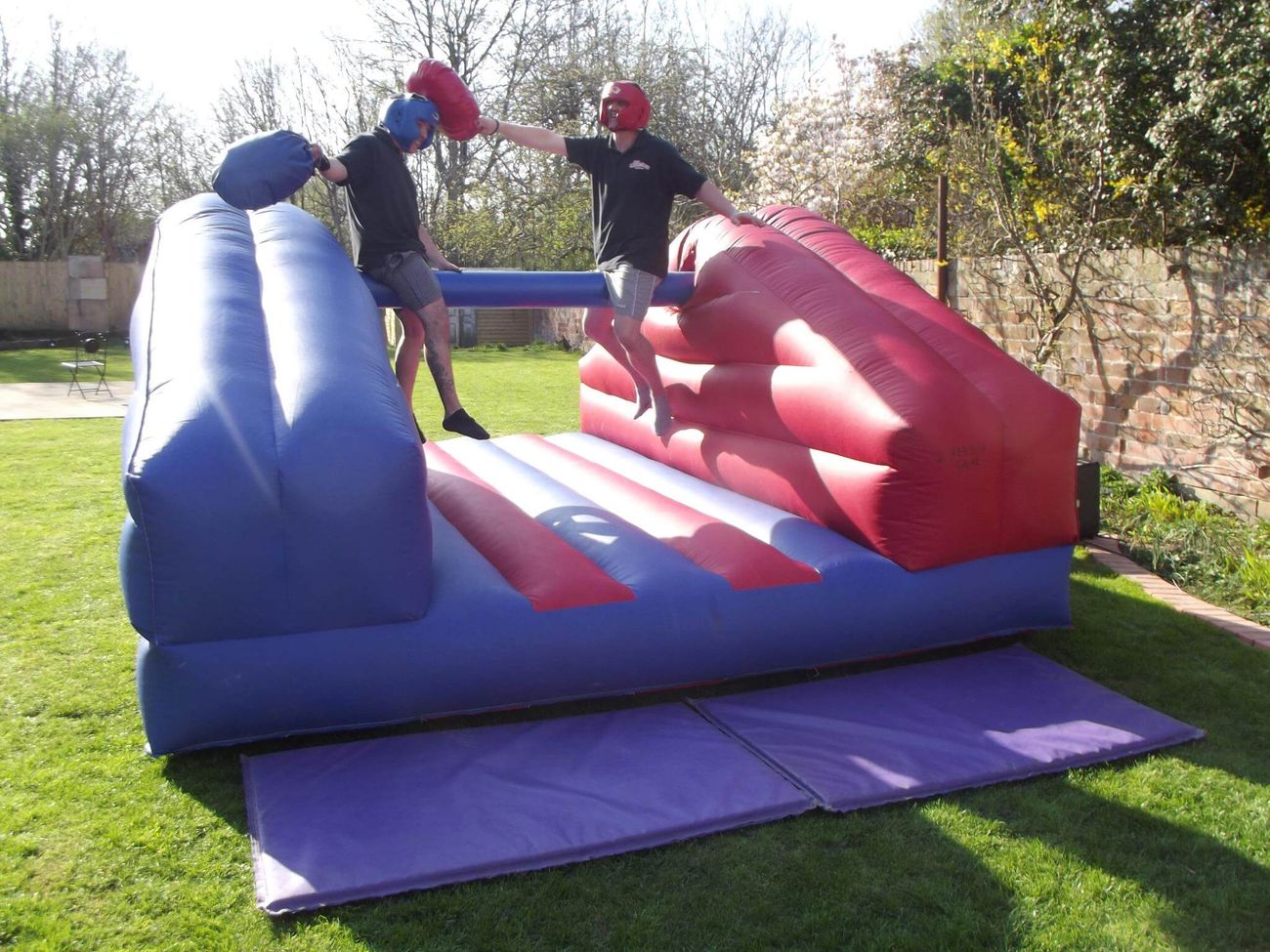 Two men in head guards astride the pole, swinging padded pillows at each other over the inflatable bed