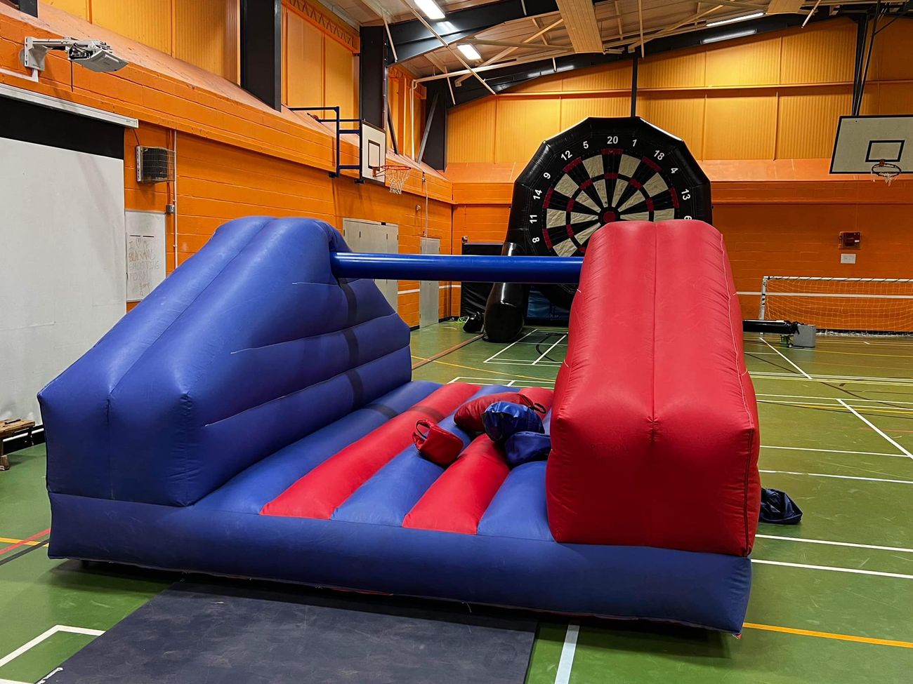 Pole jousting inflatable with padded pillows on the bed, set up in a sports hall with a giant football dartboard behind