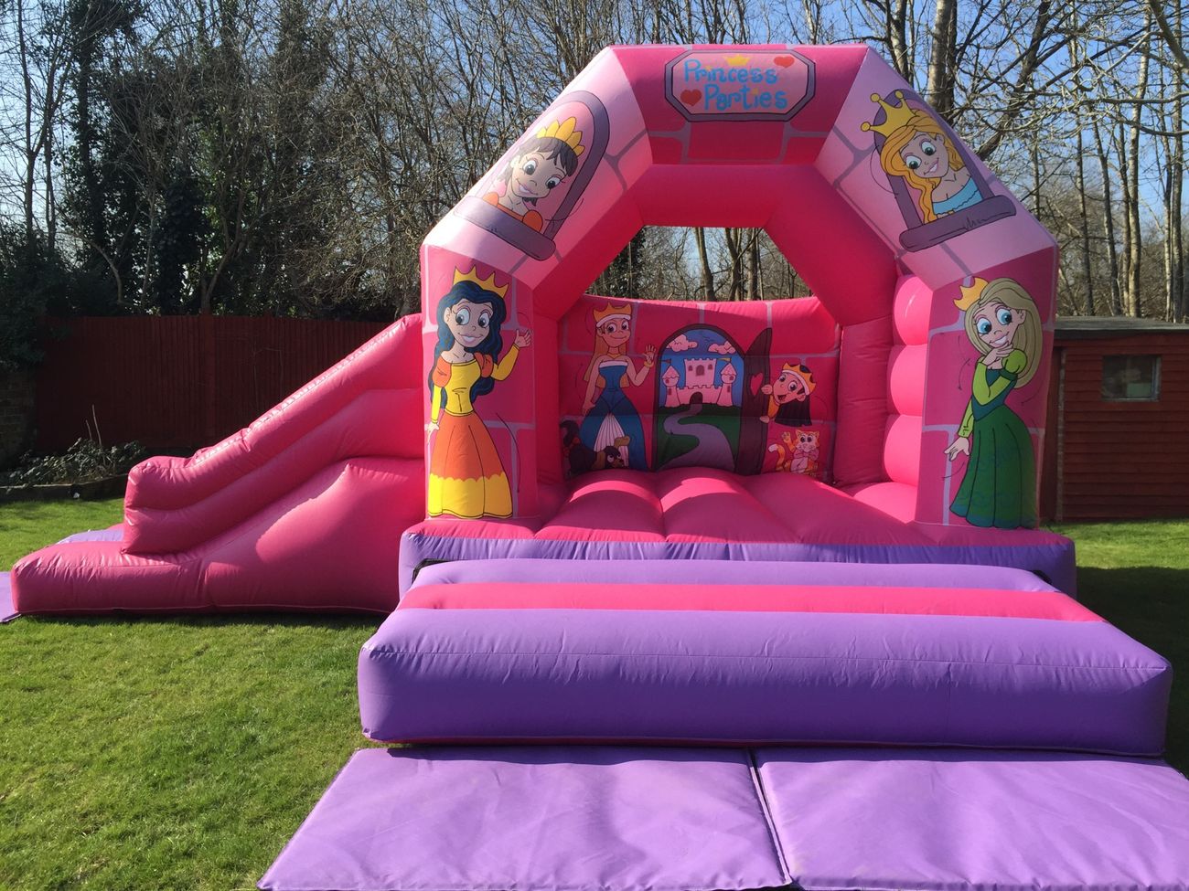 Pink and purple princess bouncy castle with a side slide and crash mats on grass