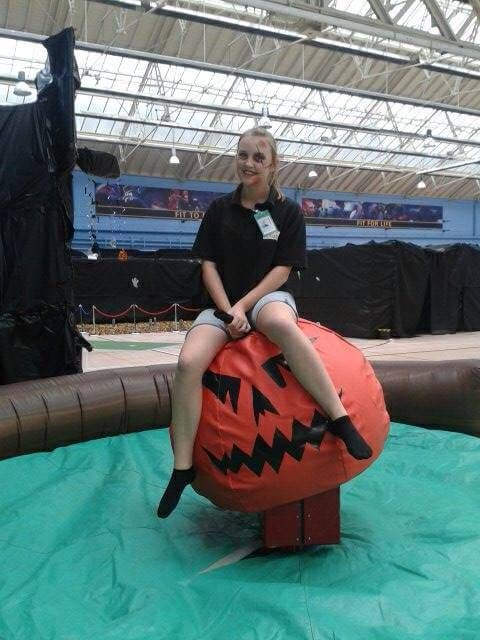Rider in Halloween face paint sitting on the carved orange rodeo pumpkin in an indoor venue