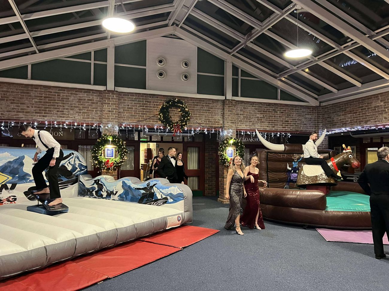 Christmas party venue with a man riding the rodeo reindeer on the right and another on a snowboard simulator on the left, guests in evening wear between