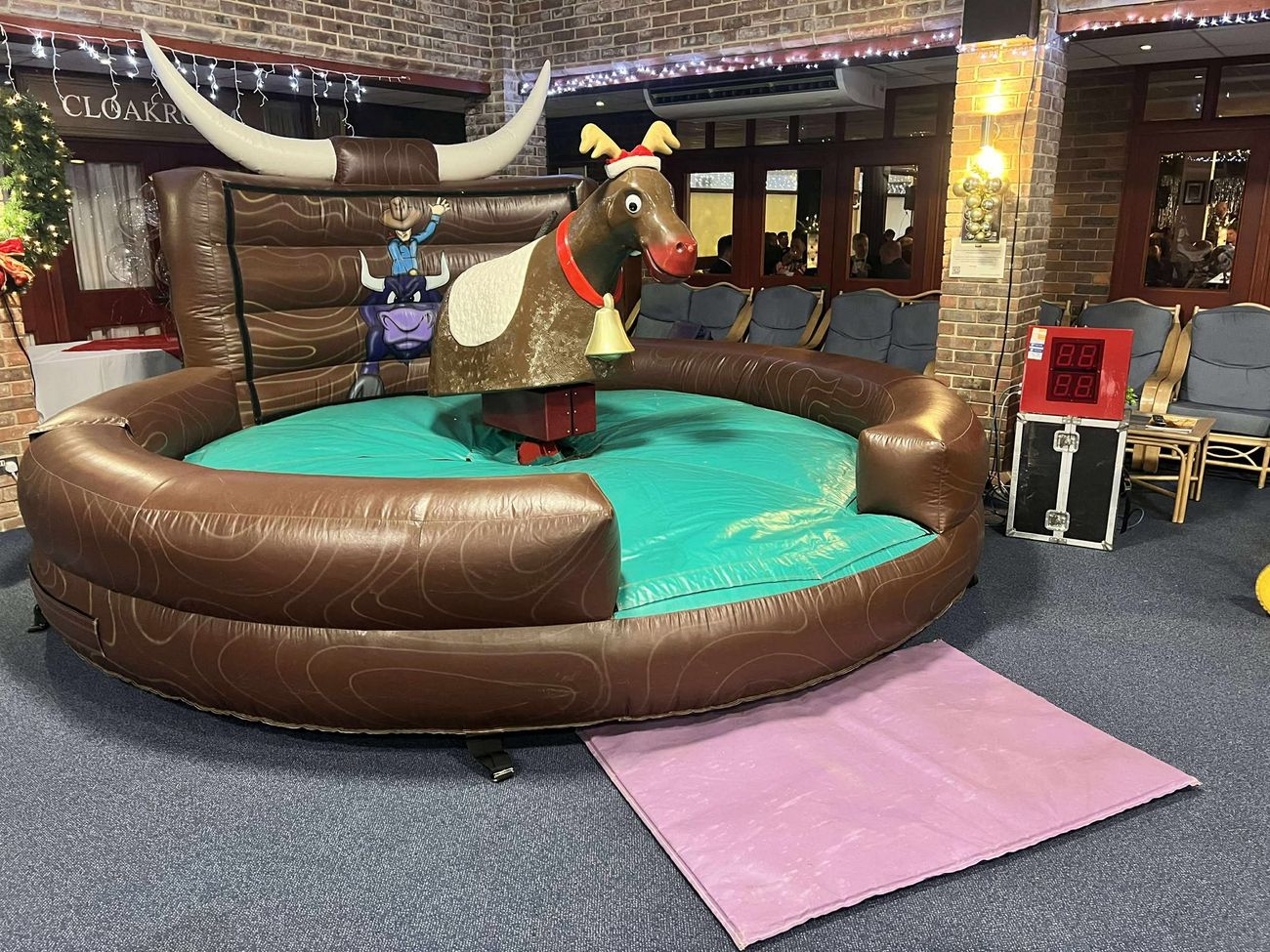 Rodeo reindeer with antlers, Santa hat and bell on a round brown inflatable bed with green centre, set up in a brick-walled venue with a timer unit