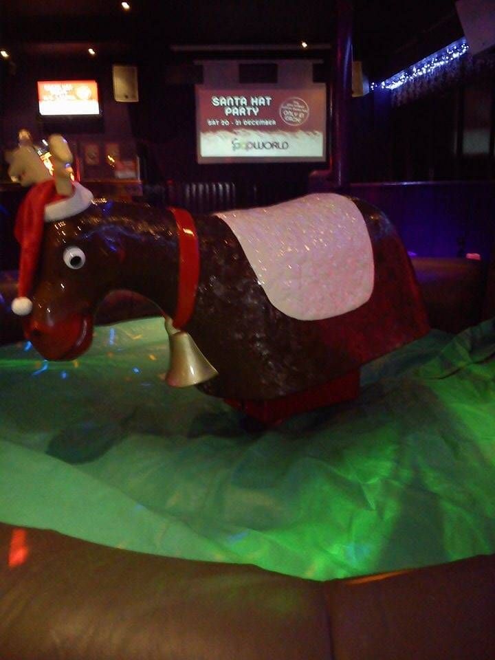 Close-up of the mechanical rodeo reindeer with red Santa hat and neck bell on a green inflatable bed at a club, Santa Hat Party screen behind