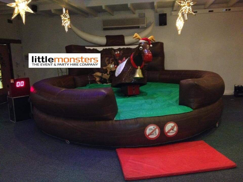 Rodeo reindeer with Santa hat and neck bell on a green and brown inflatable bed in a hall, red crash mat and digital timer alongside