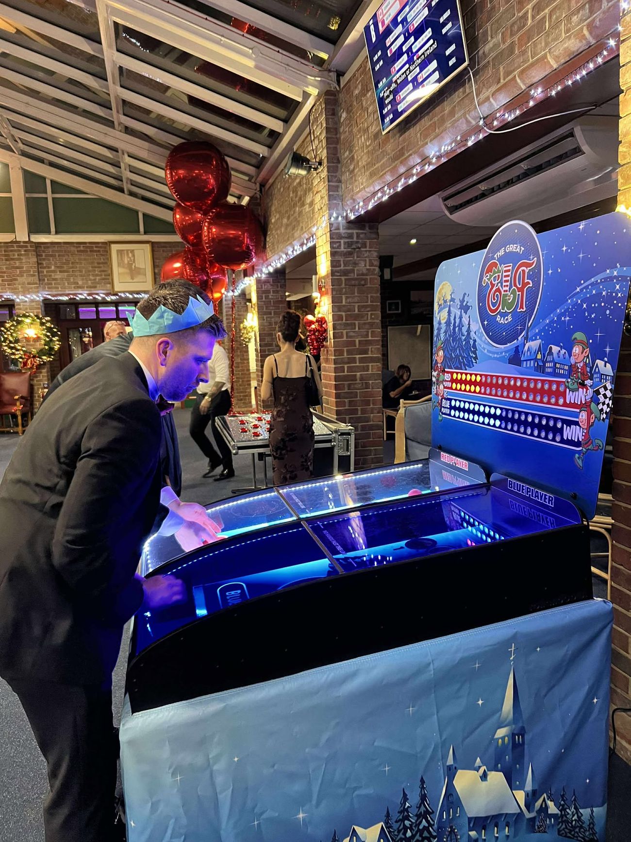 Man in a paper party hat playing the Elf Racing Derby at a Christmas party with fairy lights