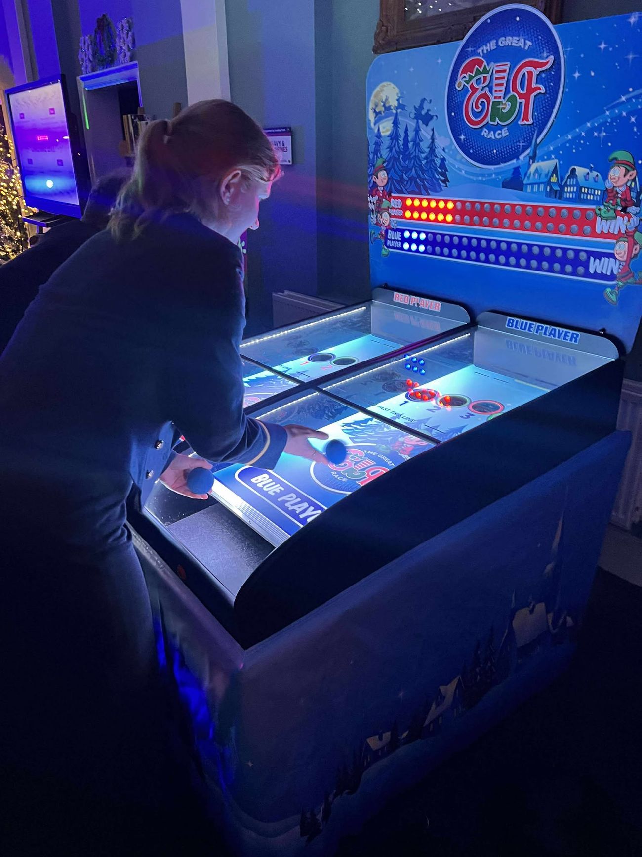 A guest rolling a ball up the blue lane of the lit elf racing derby at an evening party