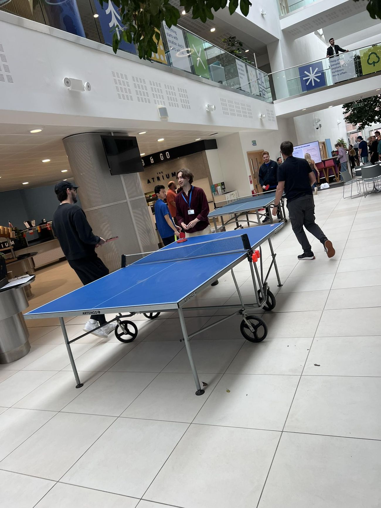 Blue table tennis table set up in a bright indoor atrium with players holding bats