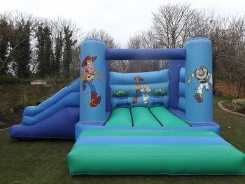 Blue Toy Story bouncy castle with Woody and Buzz artwork and a side slide on the left, set up on a lawn